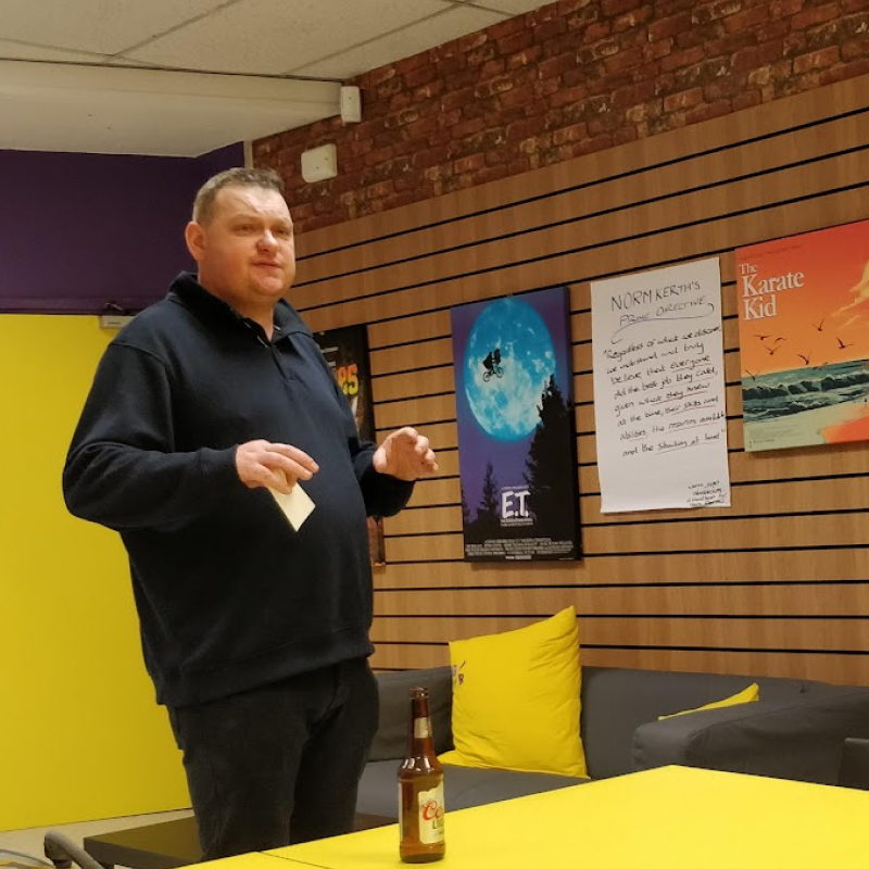 A man standing indoors gives a presentation in a lounge area decorated with movie posters and bright yellow furniture.