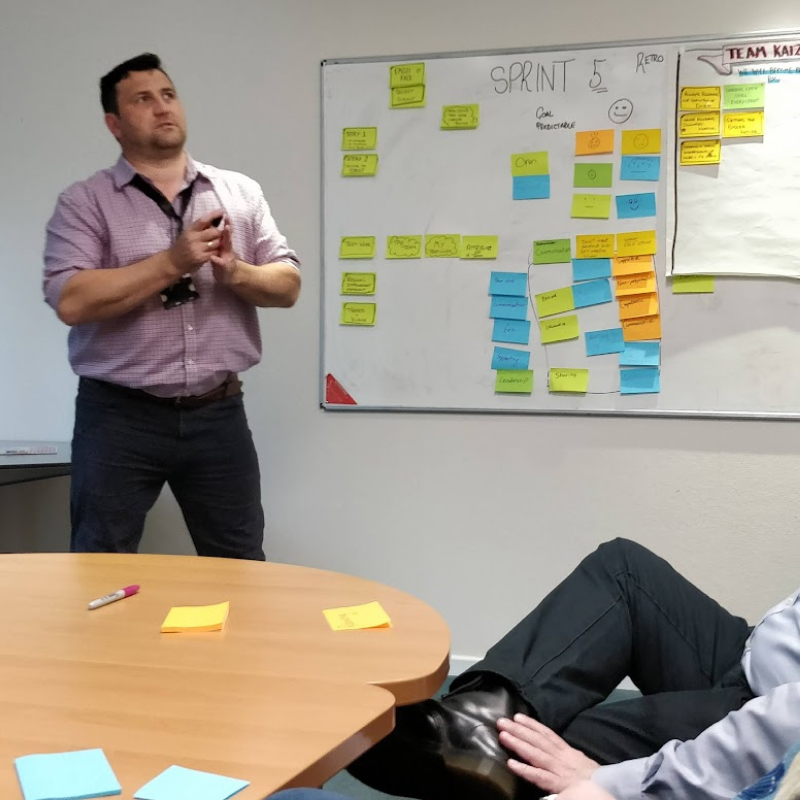 A man presents in a meeting room beside a whiteboard covered with colorful sticky notes and sprint planning details.