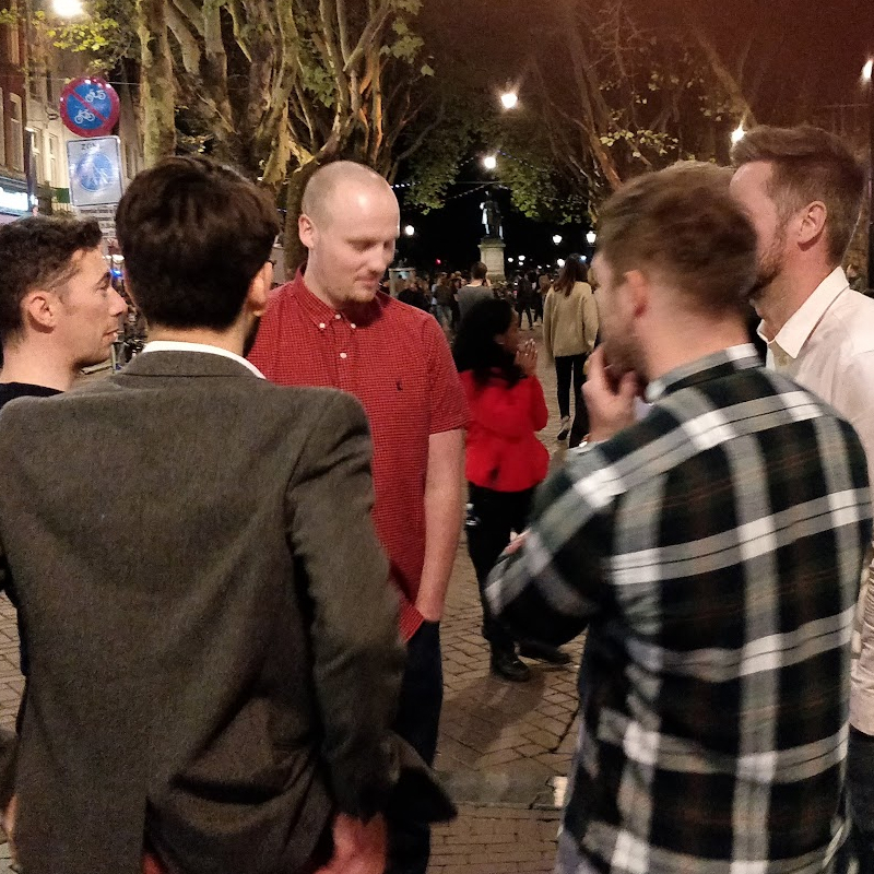 Several men stand talking together on a busy city street at night lined with trees and streetlights.